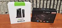 Photo showing Nvidia Shield 4K HDR Android TV box still new in box and unopened TV box marked 'TV Box'. Both boxes are placed on a wooden table outdoors.