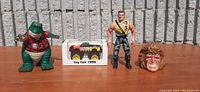 Wide shot of all four toys: dinosaur figure, Tonka truck box, Terminator 2 figure, and Warrior squirt head.