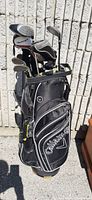 Full view of black Callaway golf bag filled with assorted golf clubs against cement wall outdoors.