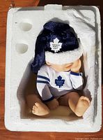 Porcelain baby doll dressed in Toronto Maple Leafs outfit seated inside Styrofoam packaging.