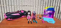 Draculaura car, two Monster High dolls (Draculaura and Catty Noir), and small pink and blue playhouse with accessories arranged on a wooden surface outdoors.