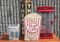Full display of all items in the lot against a gray brick wall: Sunbeam popcorn maker, Stokes popcorn bowls box, metal theater sign, and wooden popcorn sign.