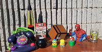 Photo showing entire lot including lava lamp, Nickelodeon purple phone, Minecraft puzzle, Spiderman bank figure, Simpsons and Mario figures on table outside against brick wall