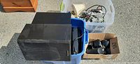 Two large black Sony speakers sitting on a blue bin filled with cords and cables, also nearby a white bin with more cords.