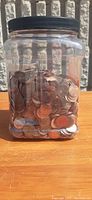Front view of a large clear plastic jar filled with numerous USA pennies showing varied copper hues and wear.