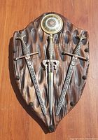 Front view of vintage wooden shield-shaped crest plaque with three miniature swords mounted on it, showing full design and details of plaque and swords.