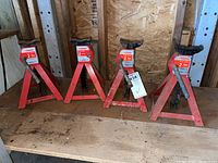 Four Motomaster red axle stands on wooden shelf, showing triangular steel bases with rust