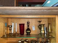 Photo of shelf with various brass candle holders, brass vases, a red glass vase and a hammered silver-tone pitcher.