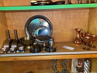 Shelf with copper decanter, hammered goblets, silver-plated trays, small bowls, coasters, and candle holders.