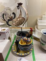Photo of large metal rooster decor with beige, brown, and black finish standing on a black base, surrounded by stacked ceramic bowls and plates, and a small ceramic rooster figurine.