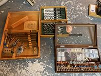 Wooden box containing multiple torch attachments with tool placement markings, next to two boxes with large drill bits and a Dremmel kit.