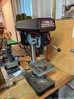 Frontal view showing the brand label on top 'DELTA 8' BASIC DRILL PRESS' with some sanding dust on the worktable and base, drill chuck visible, mounted on a workbench in a workshop.