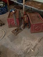 Hydraulic floor jack, bumper jack, red steel vehicle ramps, and metal block are positioned on garage floor showing signs of use and rust.