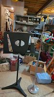 Photo showing the black metal music stand with adjustable tripod base along with silver adjustable microphone stand with round base in a cluttered room.
