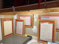 Nine framed technical drawings resting against a wall and on a table, displaying different mechanical and geometric designs with wooden frames and colored corner tapes.