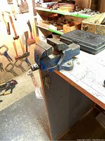 Front and side angle view of the small blue vise mounted on the edge of a workbench in a basement workshop. Displays vise jaws and handle.