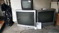 Three CRT televisions stacked and aligned: small Sanyo black model on top, large silver Signature 2000 in front center, medium silver Insignia to right side