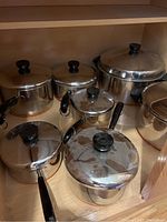 Seven stainless steel pots with lids and black handles placed on a wooden shelf inside a cabinet, showing varying sizes.