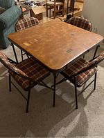 View of the square card table and four upholstered folding chairs arranged on carpeted floor, showing brown faux woodgrain top with taped holes and black metal frames.