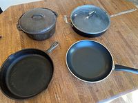 Photo showing the four pans on a wooden surface: a cast iron Dutch oven with lid, a cast iron skillet, and two nonstick pans including an emerald green pan with lid.