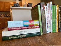 Photo showing a stack of various cookbooks including titles such as 'Alton Brown's Kitchen User Manual', 'Cooking with Love' by Carla Hall, and 'The Zuni Cafe Cookbook'.