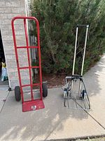 View of two dollies on pavement: one heavy-duty red dolly with large wheels, and one lightweight folding hand truck dolly with metal frame and small wheels.