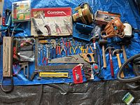 Overall layout of assorted hand tools including saws, pliers, hammers, clamps, drills, and small hardware on tarp.