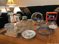 Overview of assorted glass dishes, canisters, ceramic figurines, and decorative plates arranged on a wooden table in living room setting.