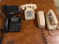 Photo showing four handheld phones and one answering machine all placed on brown surface. Phones vary in size, design, and color mostly beige and black.