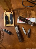 Photo showing six Leatherman multi-tools, two leather sheaths, a black watch band, and a wooden storage box.