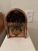 Front view of copper-toned vintage novelty music box featuring decorative scroll work and etched design resembling a jukebox or radio.
