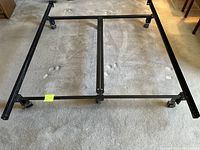 Large black metal queen bed frame laid flat on carpet, showing entire rectangular frame and wheel attachments at corners and center support.