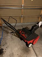 Full view of red and black MTD Yard Machine snowblower showing handle and white wheels on cement floor.