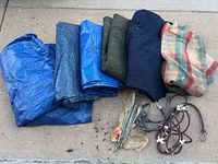 Lot showing three blue tarps, three moving blankets (dark blue, olive green, plaid), bungee cords, and rusted metal stakes on concrete surface
