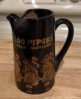 Black ceramic pitcher with gold 100 Pipers From Scotland text and three bagpiper illustrations on the front.