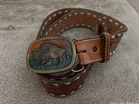 Photo showing coiled brown leather belt with white stitching and a rectangular metal belt buckle featuring a colored, raised bison image.