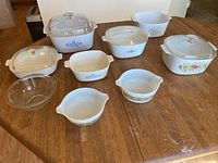 Six pieces of Corning Ware and Pyrex on wooden surface, including casserole dishes, mixing bowl, and pan handle