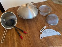 Photo showing metal wok with dome lid, metal ladle, spatula and three metal strainers plus a white plastic strainer all arranged on wooden table.