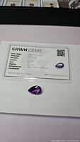 Pear shaped deep purple amethyst gemstone on certification card from GRWN GEMS showing certification date of March 18, 2023, weight 10.07 ct, color purple, clarity VVS-FL, dimensions 10.07 mm x 10.97 mm.