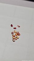 Photo showing a collection of 13 pear cut sapphires in various shades of red, orange, yellow, and pink on a white background.