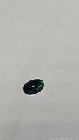 Oval cabochon cut black opal stone showing green and blue flashes under white light on a plain background.