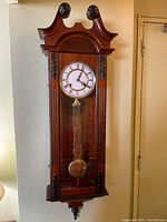 Full view of antique solid wood wall clock with glass front showing pendulum and the ornate carved wooden frame.