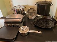 Full view of all cooking items on a black surface, showing Cuisinart electric griddle, silver pots, large black enamel pot, saucepan, strainer, and large black speckled grill pan.