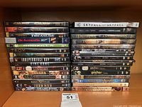 Photo showing two stacks of DVD cases on a wooden shelf with various movie titles visible.
