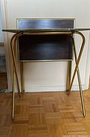 Closed and folded state of one tray table resting against a wall showing brass-colored tubular legs and dark wood veneer vertical panel.