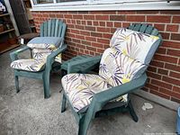 Two green plastic Muskoka chairs with floral cushions set outside against brick wall.