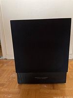 Front view of black speaker with fabric mesh covering and wood-grain lower section.