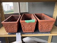 Three long rectangular terra cotta planters sitting on a shelf side by side. Each planter has embossed leaf patterns wrapping around the sides.