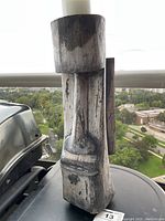 Image of weathered wooden tiki candle holder outdoors with a view of trees and buildings in background. Shows the front of the holder with carved tiki face and hollow top for candle.