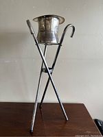 Silver-tone vintage champagne ice bucket shaped like a top hat on a tripod made from three canes seen from front left angle on wooden surface against beige wall.